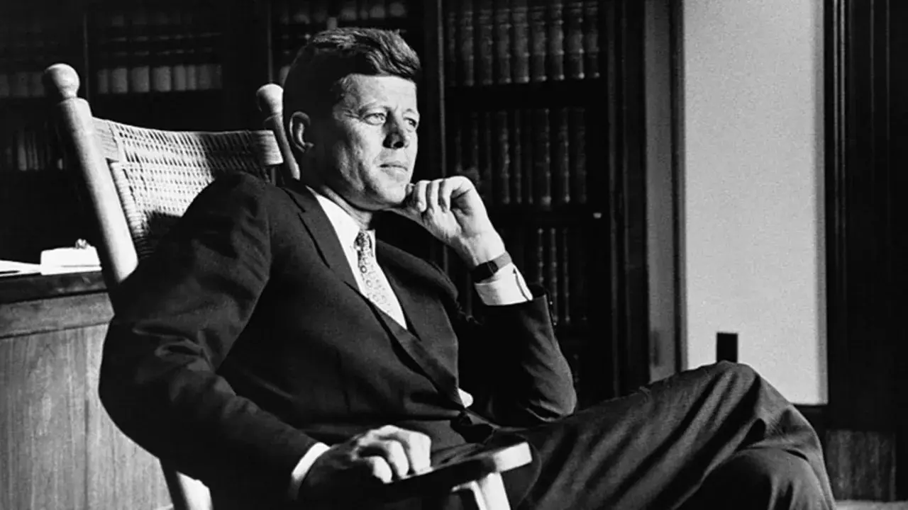 John F. Kennedy sitzt nachdenklich in einem Schaukelstuhl, seine Hand am Kinn. Trotz seiner bekannten **John F. Kennedy Krankheit** strahlt er eine ruhige Entschlossenheit aus.