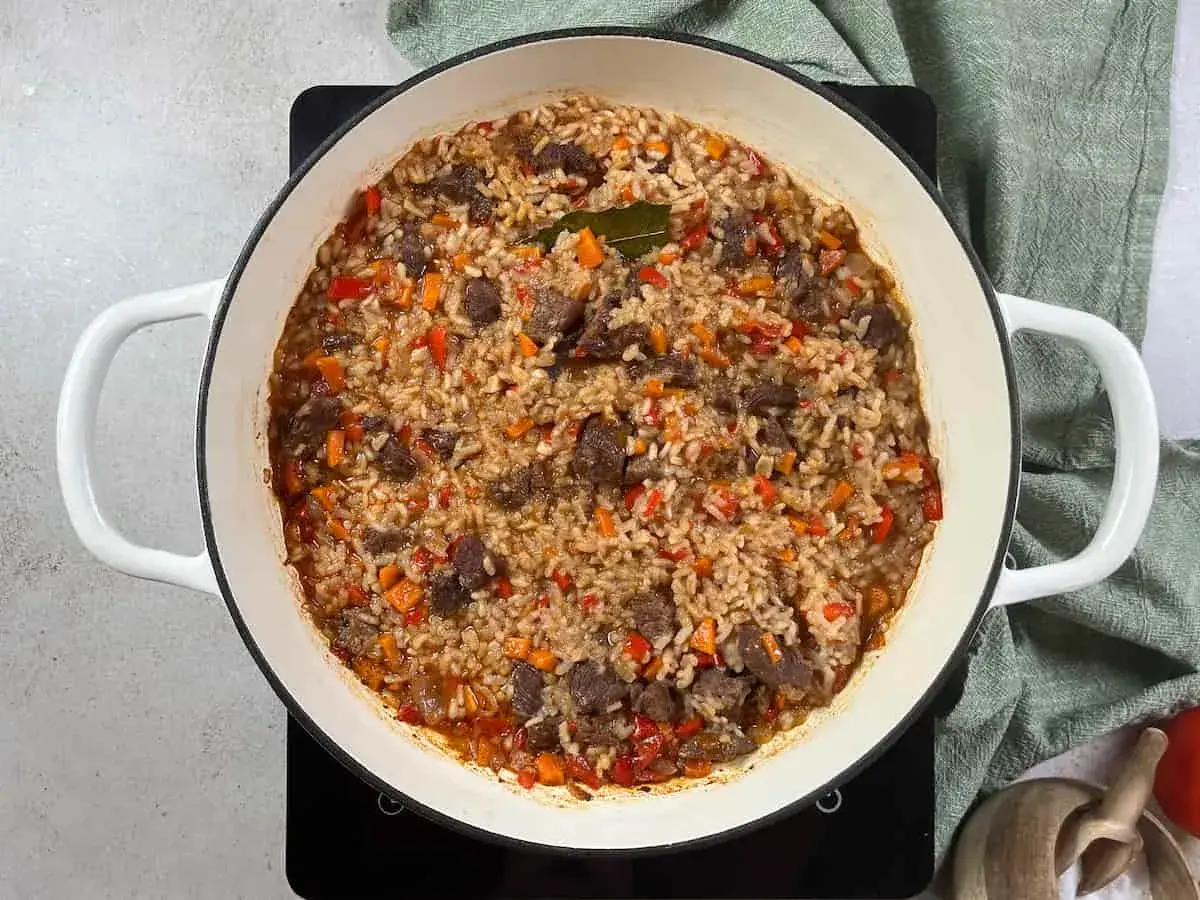 Arroz con carne y verduras, cocinado en una olla blanca. Los trozos de carne se mezclan con el arroz y pimientos rojos y zanahorias picadas.
