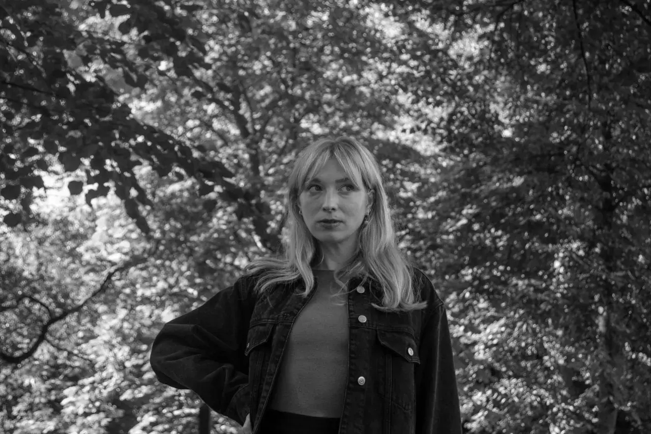 Sandra Beijer, med sitt blonda hår och blick mot horisonten, står i en skog. Hon bär en jeansjacka.