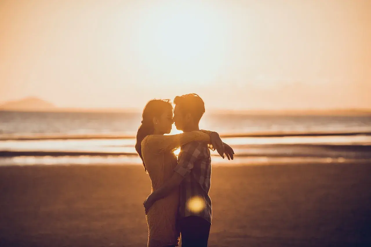 Para zakochanych na plaży o zachodzie słońca. Mężczyzna patrzy kobiecie na usta, obejmując ją.