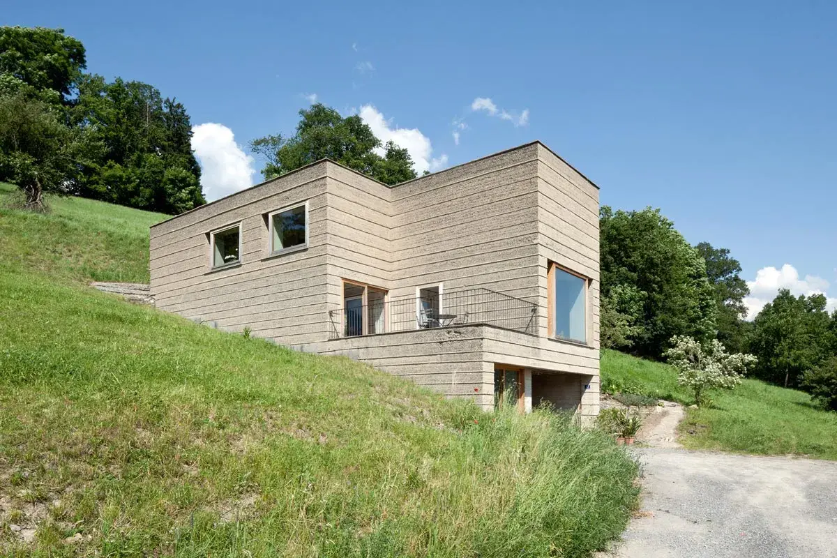 Modernes Haus mit rauer Fassade, das die natürliche Textur von **Lehm als Baustoff** zeigt. Eingebettet in eine grüne Hügellandschaft unter blauem Himmel.