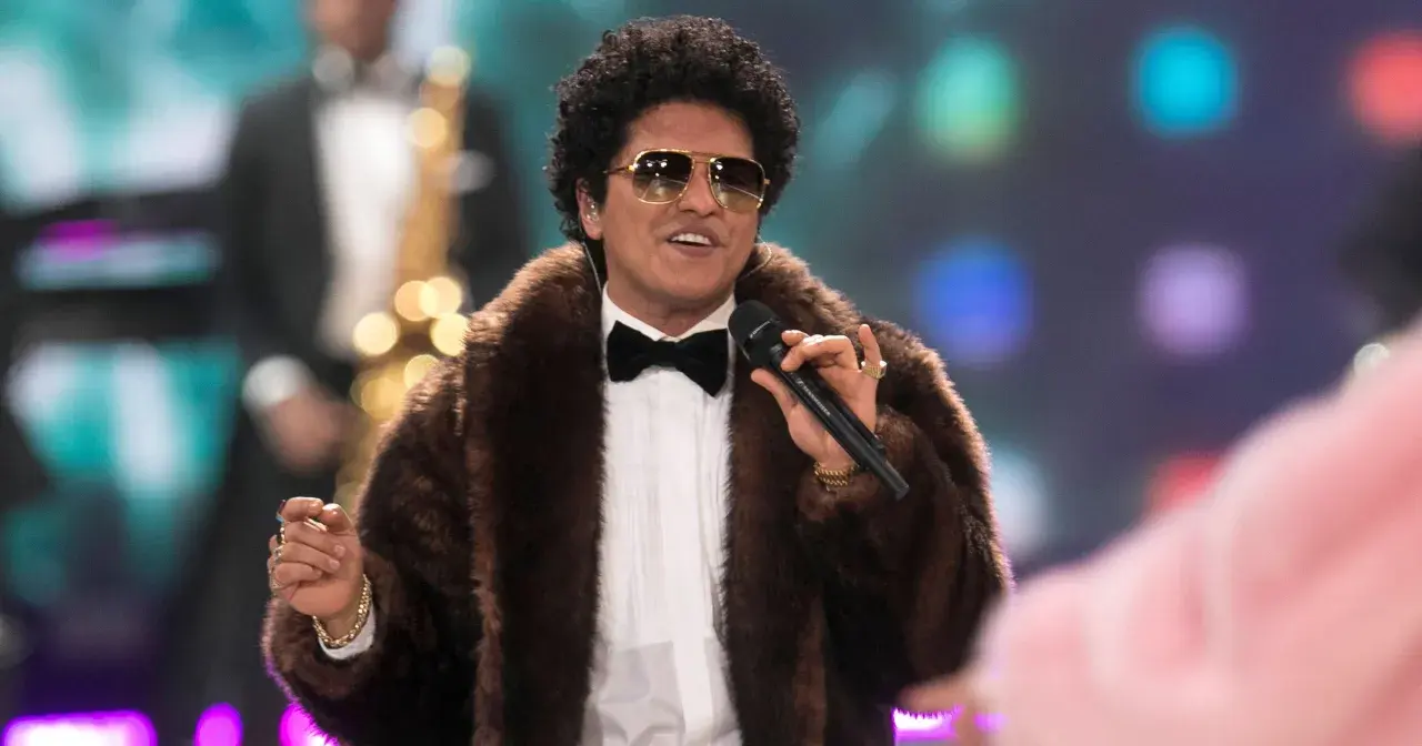 Bruno Mars w futrzanym płaszczu i okularach przeciwsłonecznych śpiewa na koncercie, trzymając mikrofon.