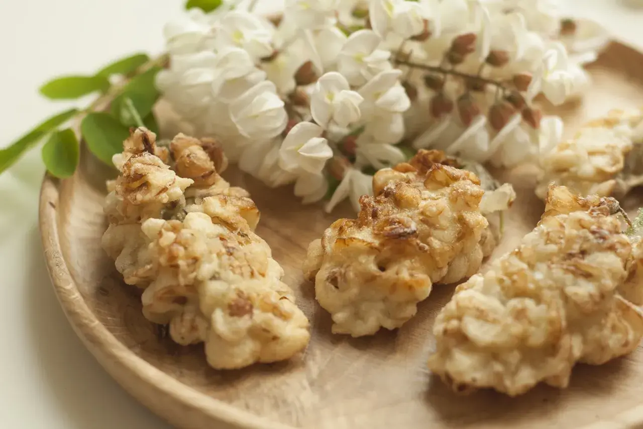 Fiori di acacia fritti dorati su un piatto di legno, con grappoli di fiori freschi sullo sfondo.