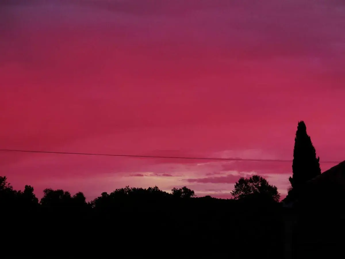 Un ciel rouge le soir, teinté de rose, surplombe une silhouette d'arbres et d'une maison.