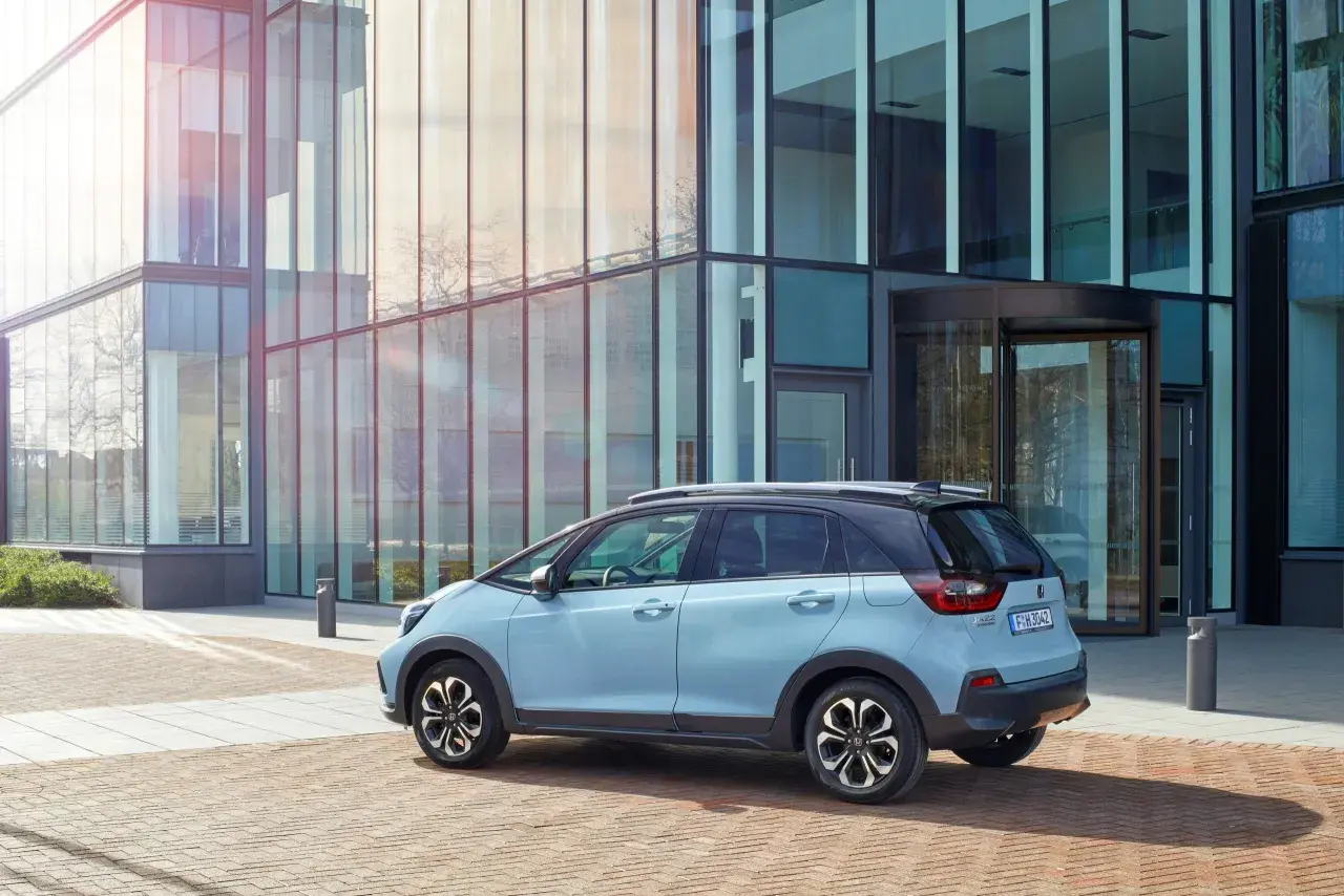 Jasnoniebieski Honda Jazz 4 stoi przed nowoczesnym budynkiem z przeszkloną fasadą.