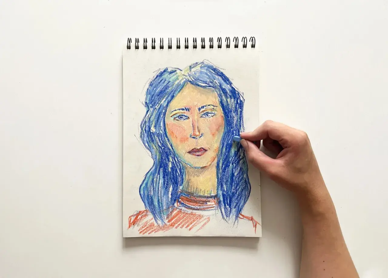 Portret kobiety z niebieskimi włosami, tworzony pastelami. Ręka dodaje ostatnie pociągnięcia, tworząc efekt dragana.