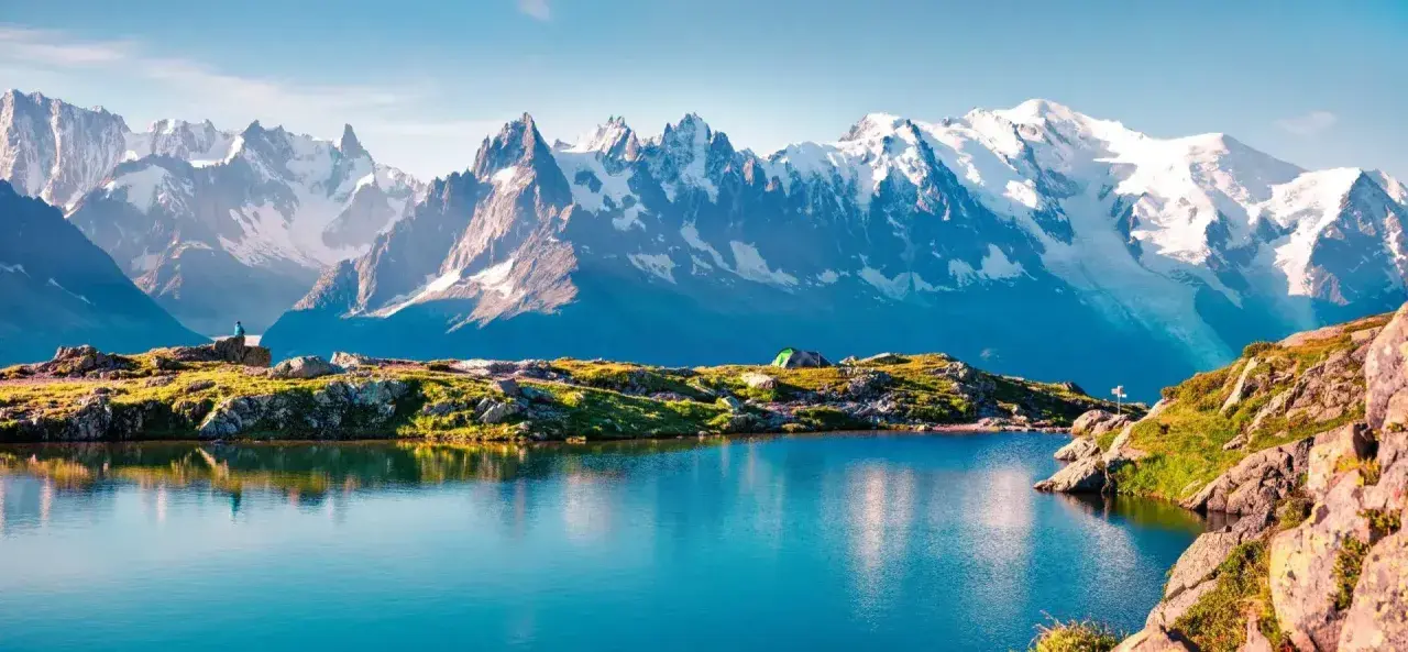 Lac alpin paisible reflétant les sommets enneigés du Mont-Blanc. Un campement discret et un randonneur contemplent ce paysage grandiose, parfait pour une randonnée tour du mont blanc.