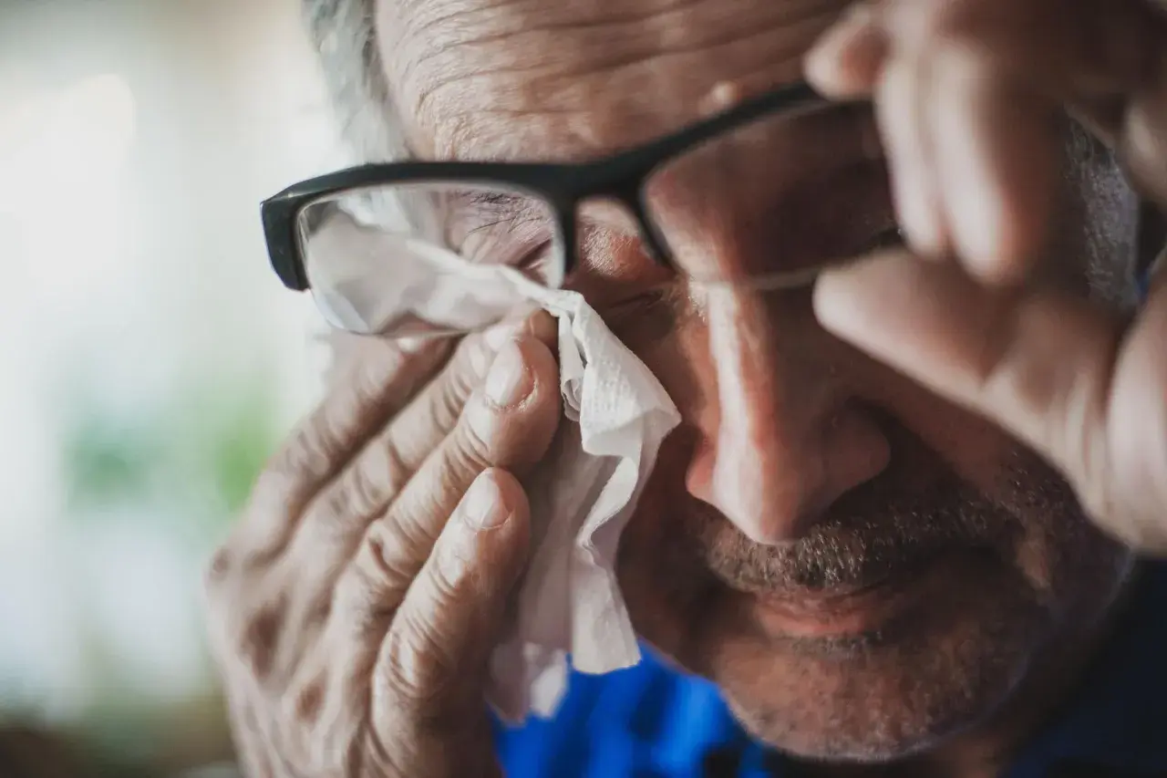 Starszy mężczyzna w okularach wyciera łzę chusteczką. Może to być objaw smutku lub reakcja alergiczna, np. na peritol.