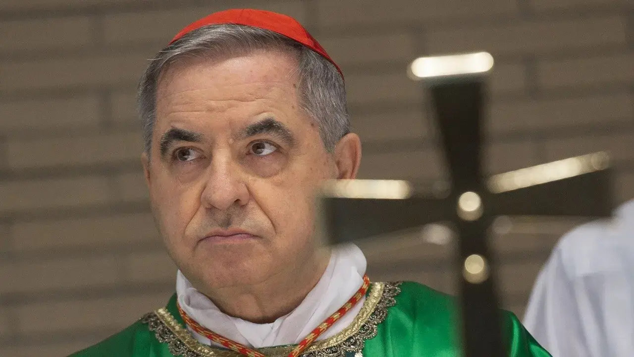 Kardinal Becciu trägt eine rote Pileolus und eine grüne Soutane. Ein Kreuz ist im Vordergrund unscharf.