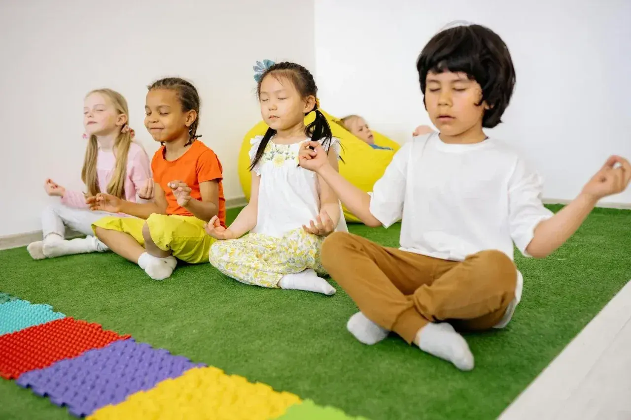 Kinder üben Achtsamkeit im Unterricht, sitzen im Schneidersitz und meditieren. Ein Junge im Vordergrund führt die Übung an.