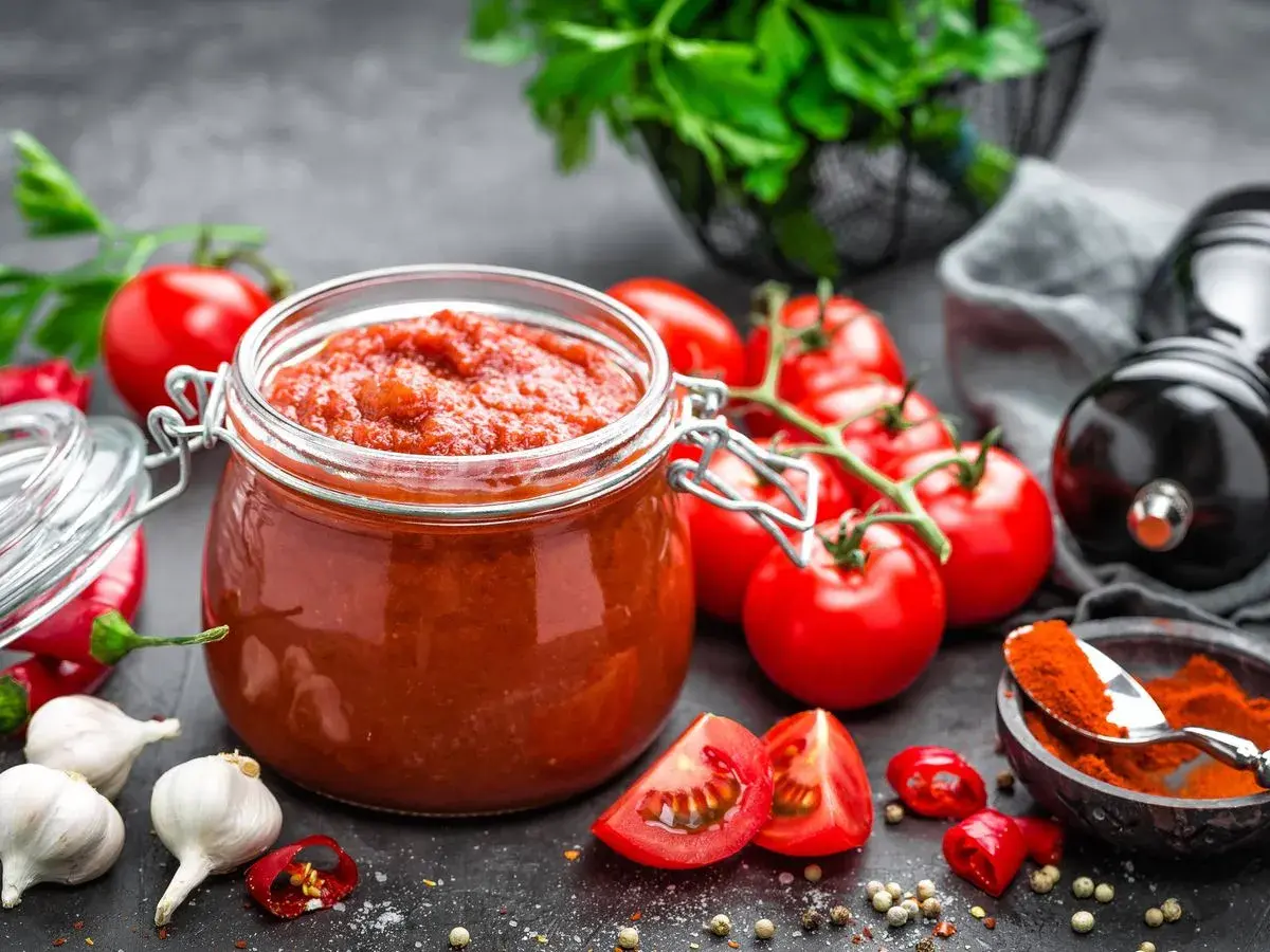 Tarro de cristal lleno de vibrante salsa de tomate casera, rodeado de tomates frescos, ajos, guindillas y especias.