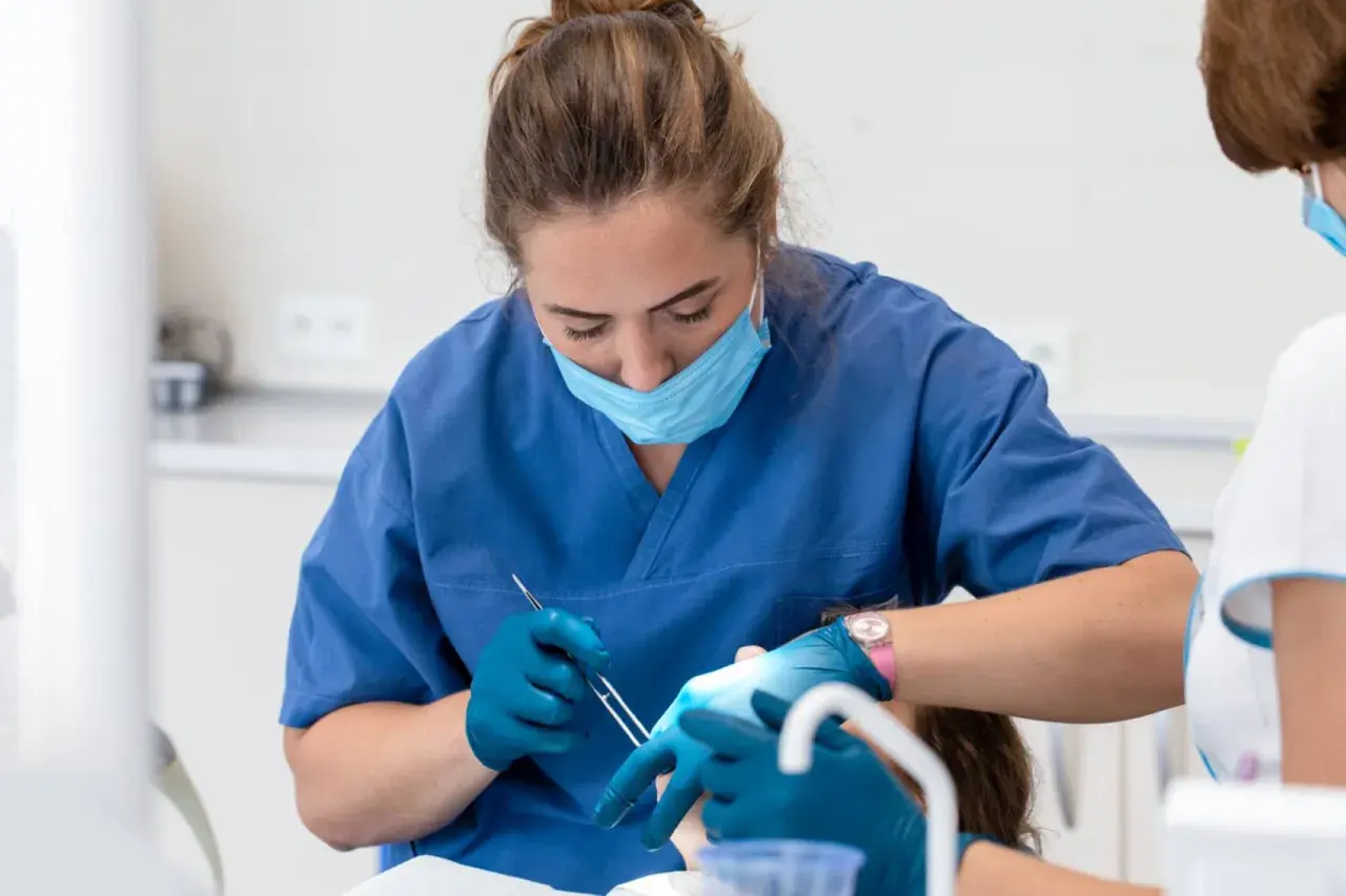 Dentystka w niebieskim fartuchu i masce zakłada opatrunek na zęba pacjentowi.