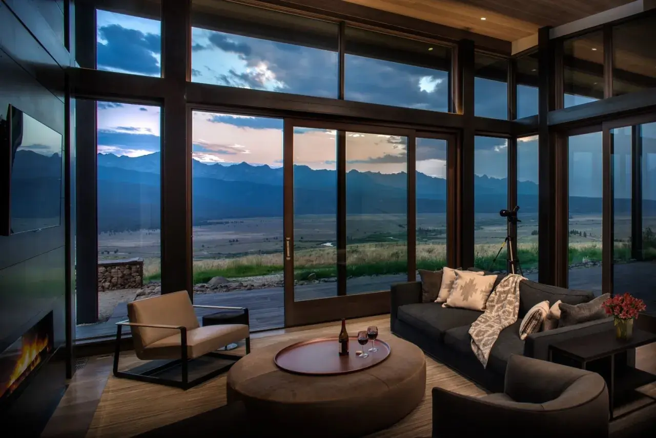Cozy living room in a modern mountain house, featuring a fireplace, plush seating, and expansive windows with a breathtaking mountain vista at dusk.