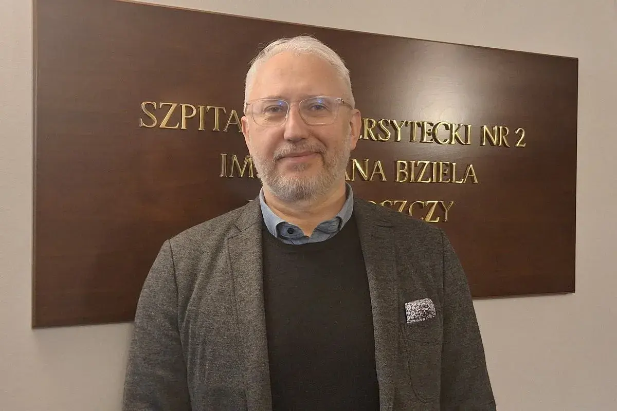 Mężczyzna w okularach i marynarce stoi przed tablicą z napisem "SZPITAL UNIWERSYTECKI NR 2 IM. JANA BIZIELA BYDGOSZCZ".