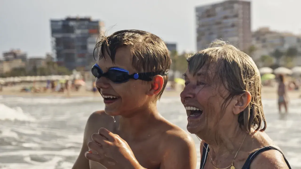 Chłopiec w goglach i starsza kobieta śmieją się na plaży. Wakacje bez zmartwień dzięki najlepszemu ubezpieczeniu turystycznemu.