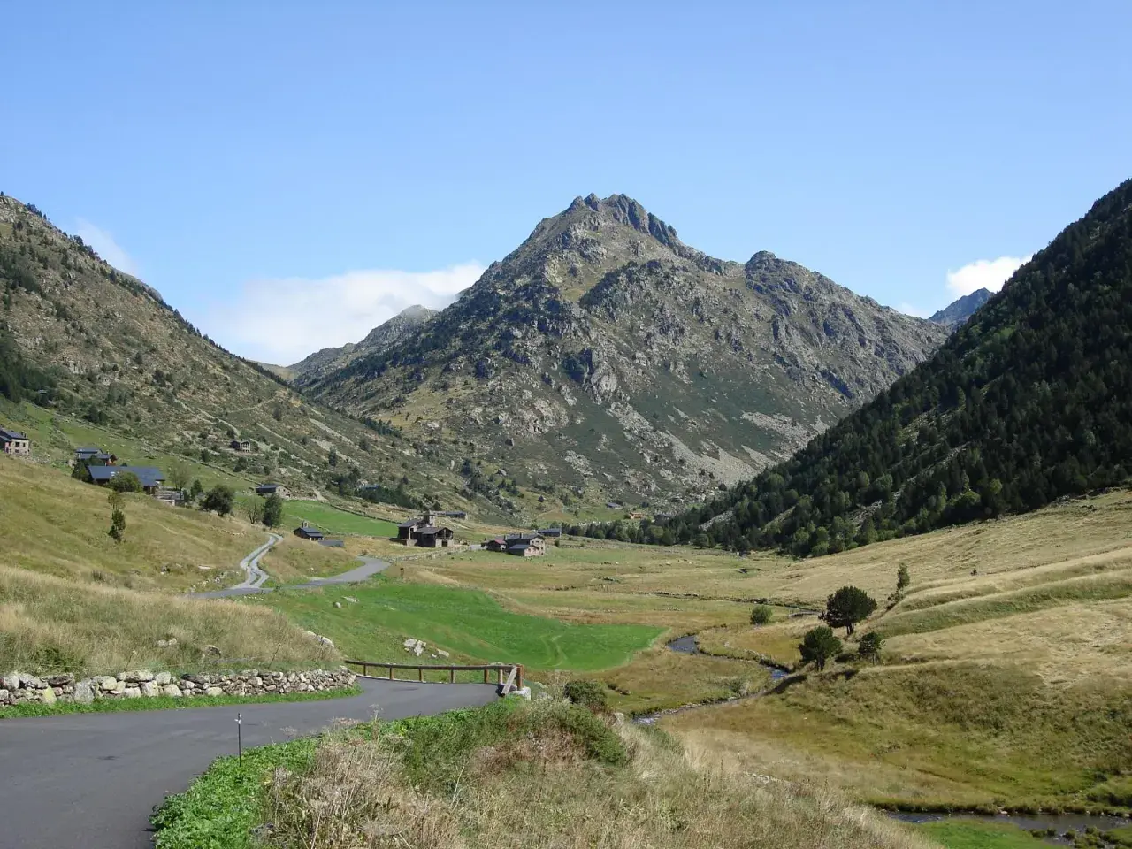 Un camino serpentea hacia el estany de Juclar, un valle andorrano con casas dispersas y un pico imponente.