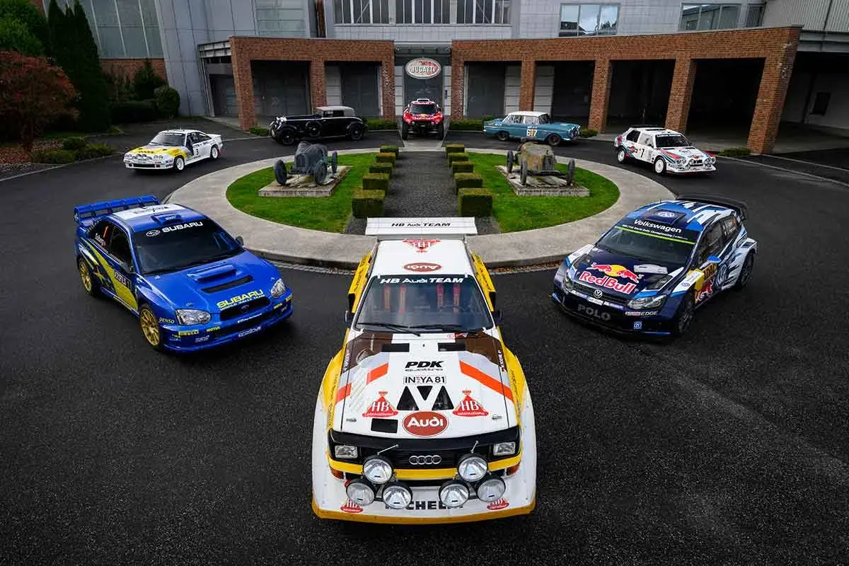 Blauer Subaru WRC Rallye-Bolide neben einem Audi Quattro und einem VW Polo. Historische Rennwagen im Hintergrund.
