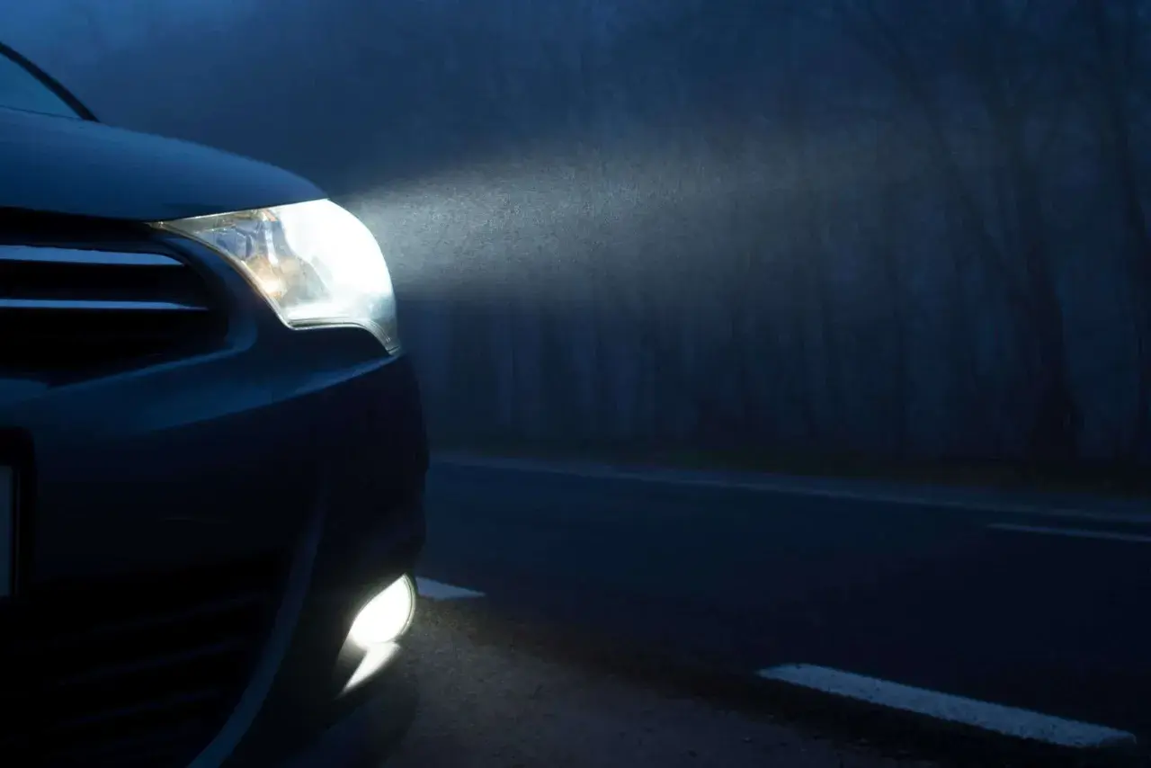 Las luces de posición de un coche iluminan la niebla en una carretera oscura, revelando el camino y los árboles a los lados.
