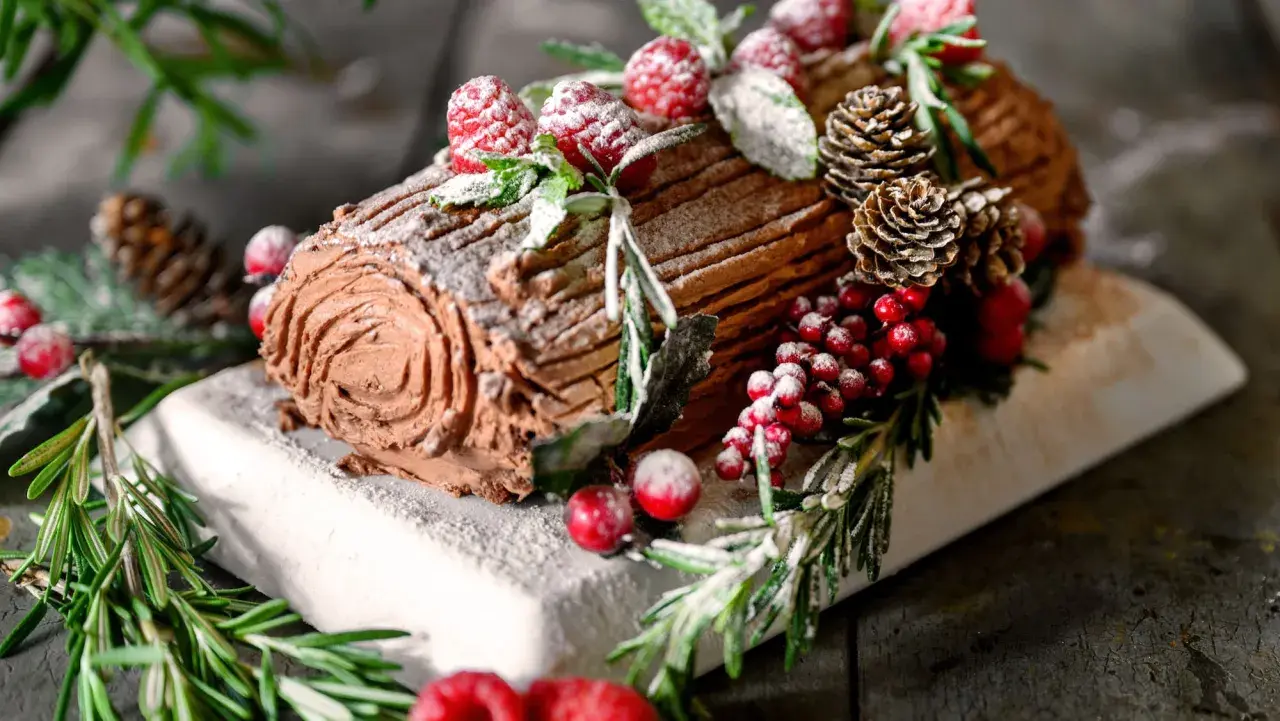Ein köstliches Bûche de Noël Rezept Original: Schokoladenrolle mit Beeren, Tannenzapfen und Rosmarin, bestäubt mit Puderzucker.