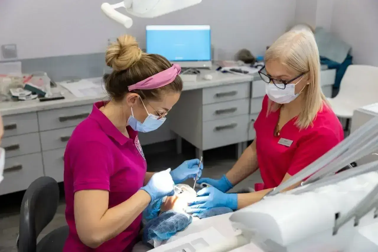Dwie dentystki w maseczkach pracują przy pacjencie. Jedna z nich podaje narzędzia, druga wykonuje zabieg, stosując lidokainę znieczulenie miejscowe.