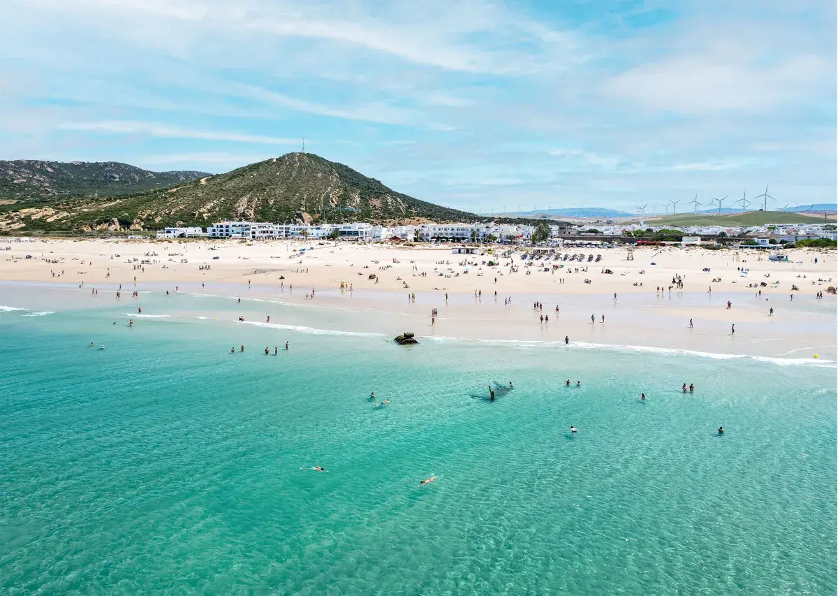 Playa de Zahara de los Atunes con gente bañándose y disfrutando del sol, con el pueblo blanco al fondo y molinos de viento en la colina.