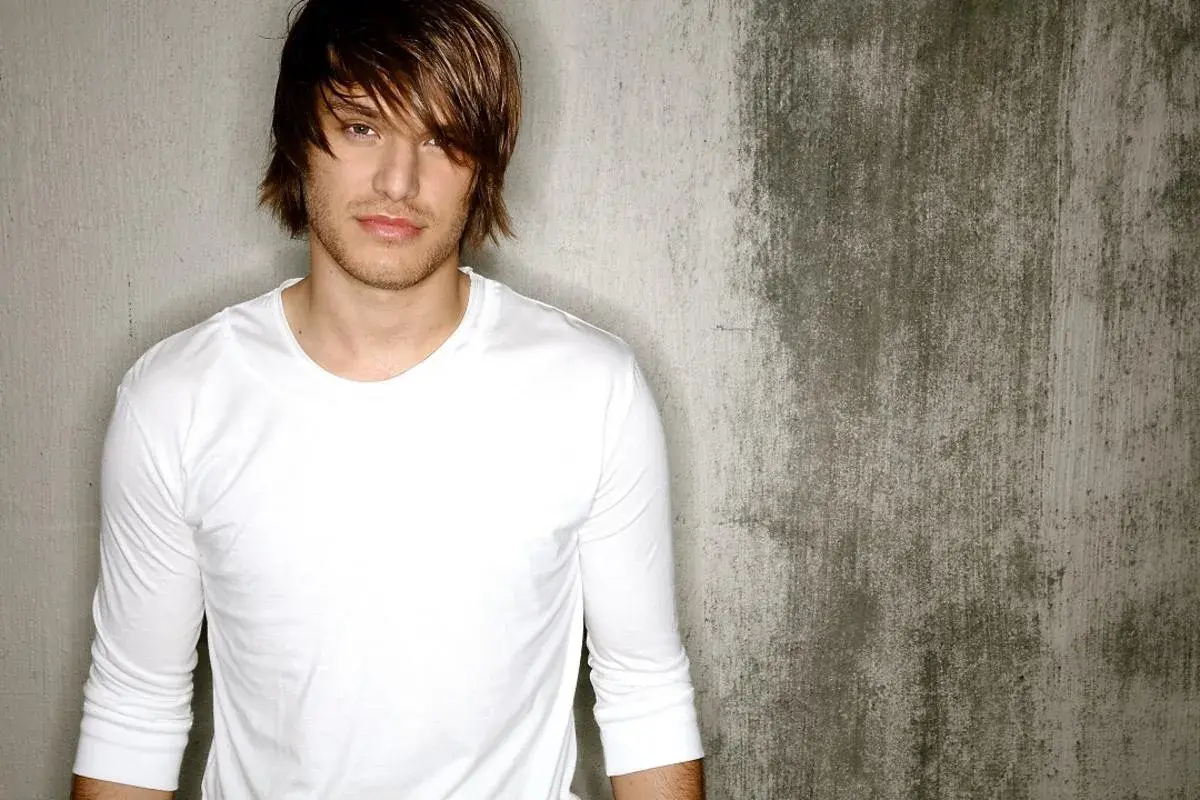 Michael Tyler, a young man with tousled brown hair and a white long-sleeved shirt, poses against a textured concrete wall.