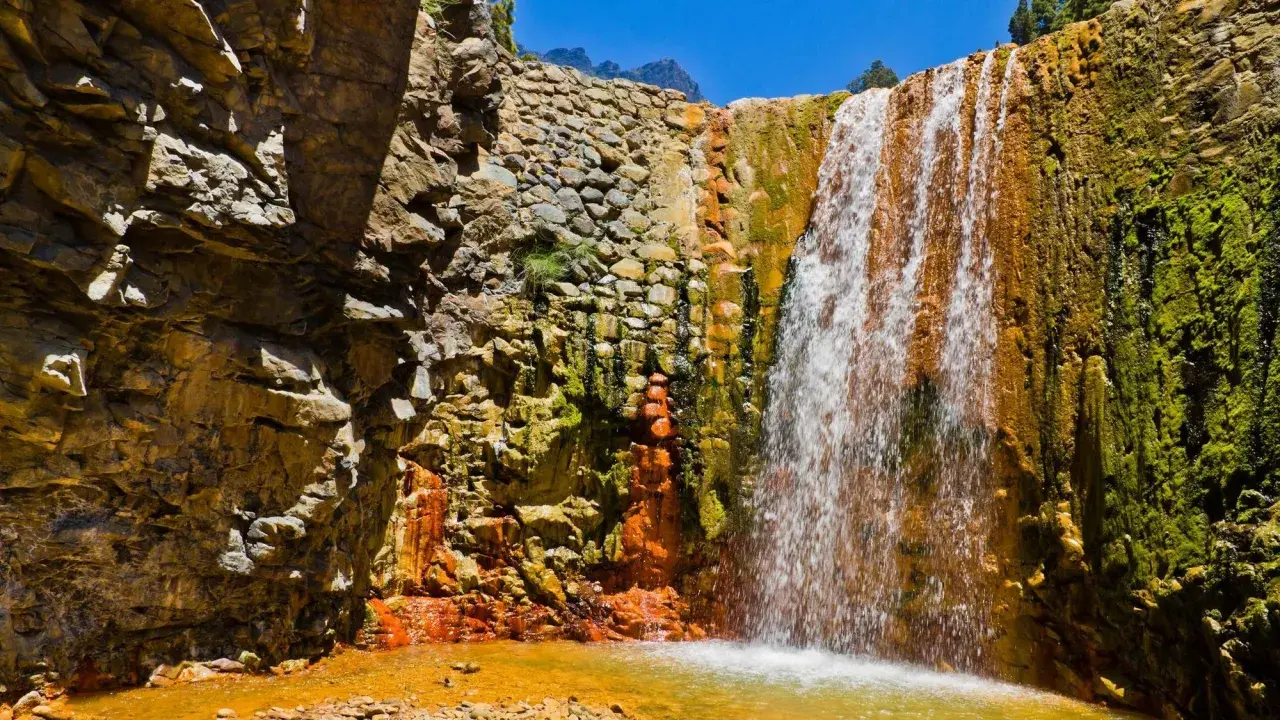 Ein Wasserfall stürzt über moosbewachsene, orangefarbene Felsen. Die Felsen sind von der Sonne beschienen, und das Wasser sammelt sich in einem goldenen Becken. Ein Stück Natur auf La Palma.