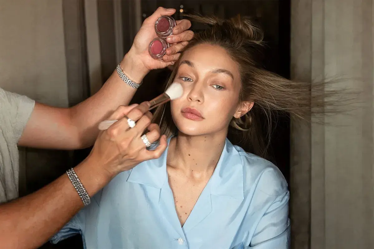 En makeupartist applicerar rouge på en kvinna som håller två små burkar med rouge. Kvinnan har sitt hår uppsatt och bär en ljusblå skjorta.