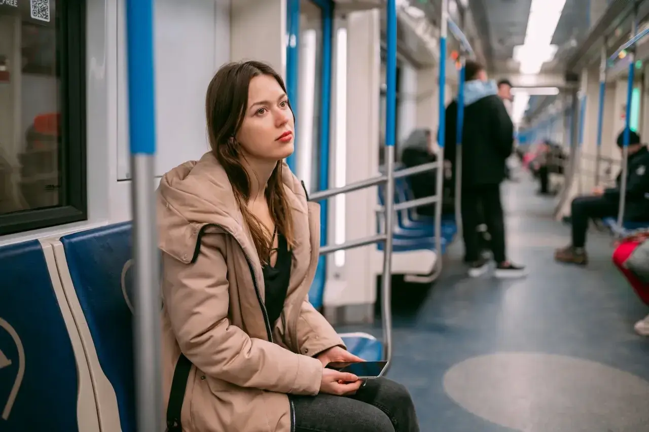 Młoda kobieta w beżowej kurtce siedzi w metrze, trzymając telefon. Jej zamyślona mina sugeruje **wypalenie zawodowe**, być może szuka ulgi lub sposobu na L4.