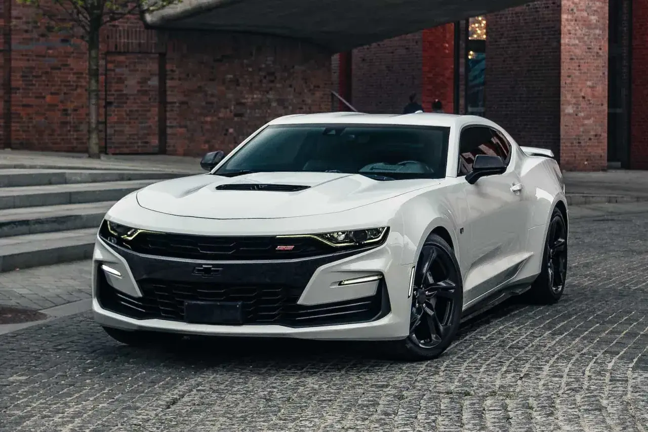 Biały Chevrolet Camaro SS z czarnymi felgami stoi na brukowanej ulicy przed ceglanym budynkiem.