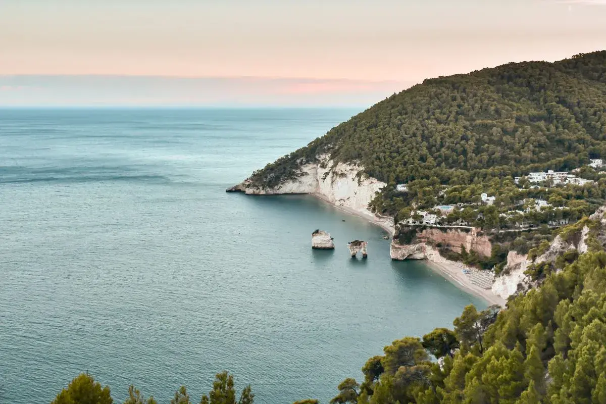 Baia con scogliere bianche e mare azzurro, perfetta per una passeggiata dell'amore. Alberi verdi e case bianche punteggiano la costa.