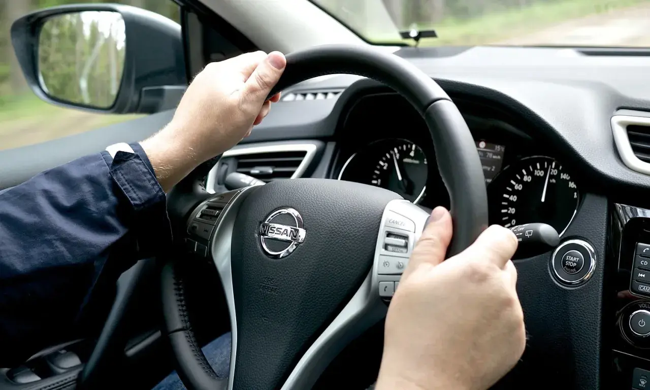 Manos al volante de un Nissan, sintiendo la vibración que indica porque vibra el volante del coche al conducir por un camino rural.