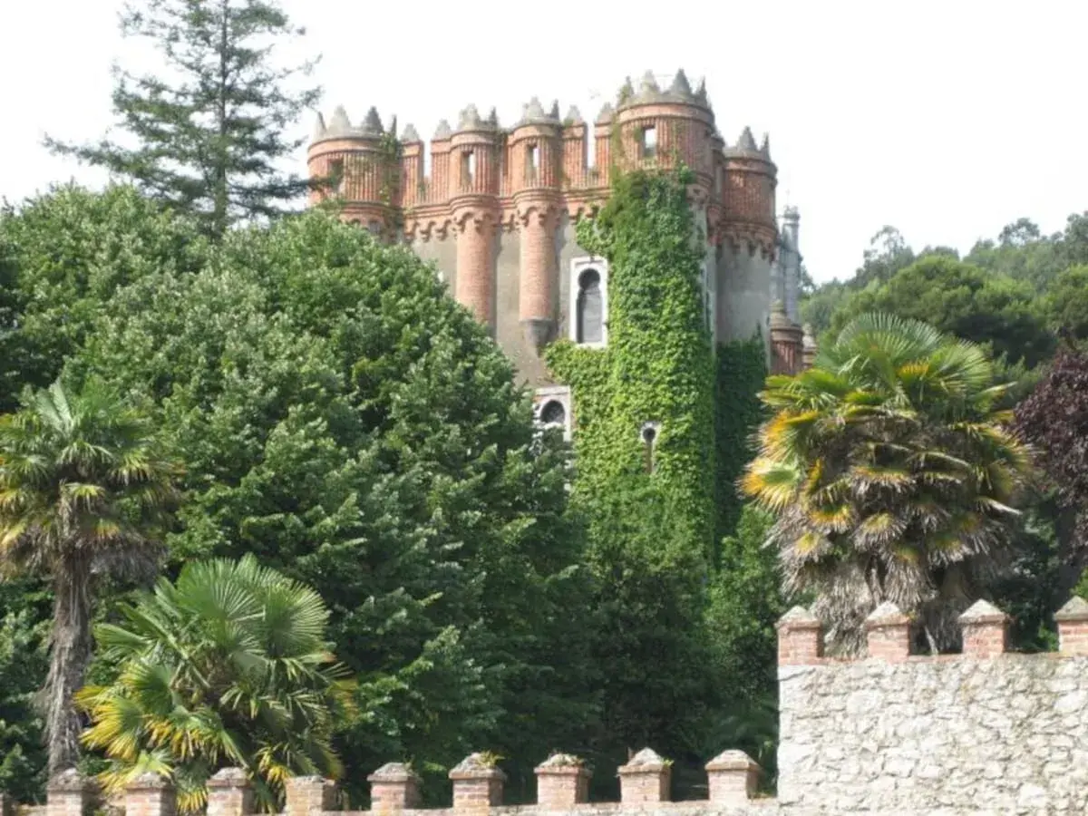 El **palacio de Ocharan**, cubierto de hiedra, se alza entre exuberante vegetación y palmeras, con un muro de piedra en primer plano.