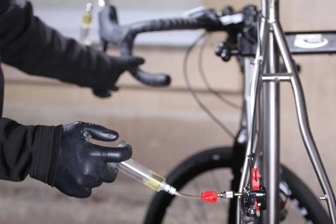 Hands in black gloves perform a bike brake bleed, following the correct brake bleed order to ensure optimal performance.