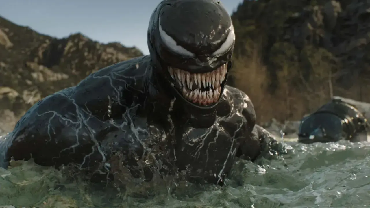 Venom émerge de l'eau, prêt pour la sortie de Venom 4. Le symbiote montre ses dents acérées dans un paysage rocheux.