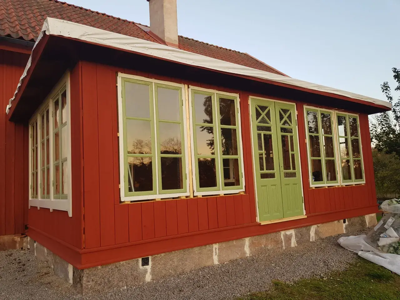 Ett rött hus med pågående arbete för att bygga glasveranda. Gröna fönster och dörrar reflekterar himlen.