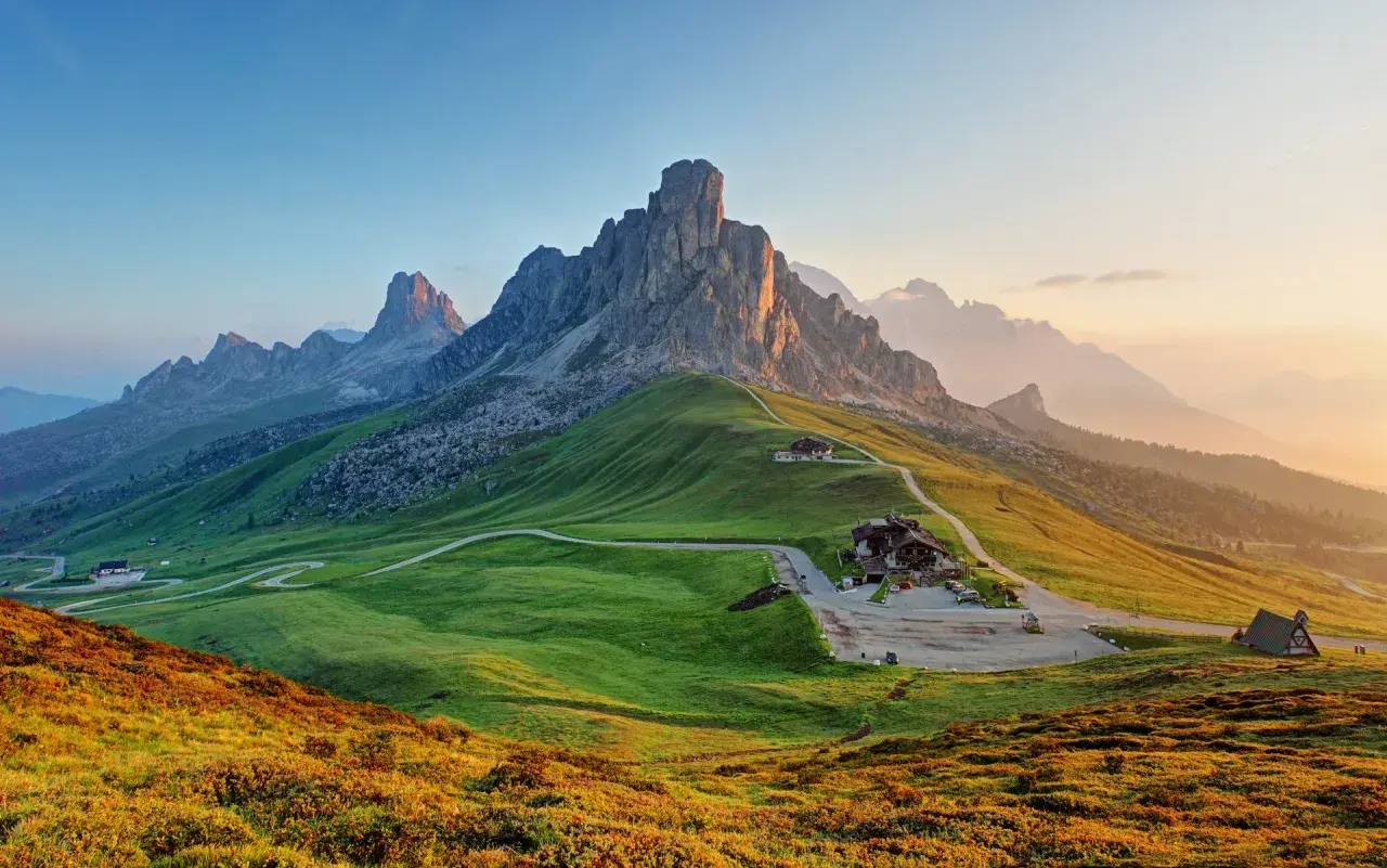 A winding road leads to a mountain lodge, perfect for hiking the Dolomites, Italy. Lush green hills meet jagged peaks under a soft sunrise.