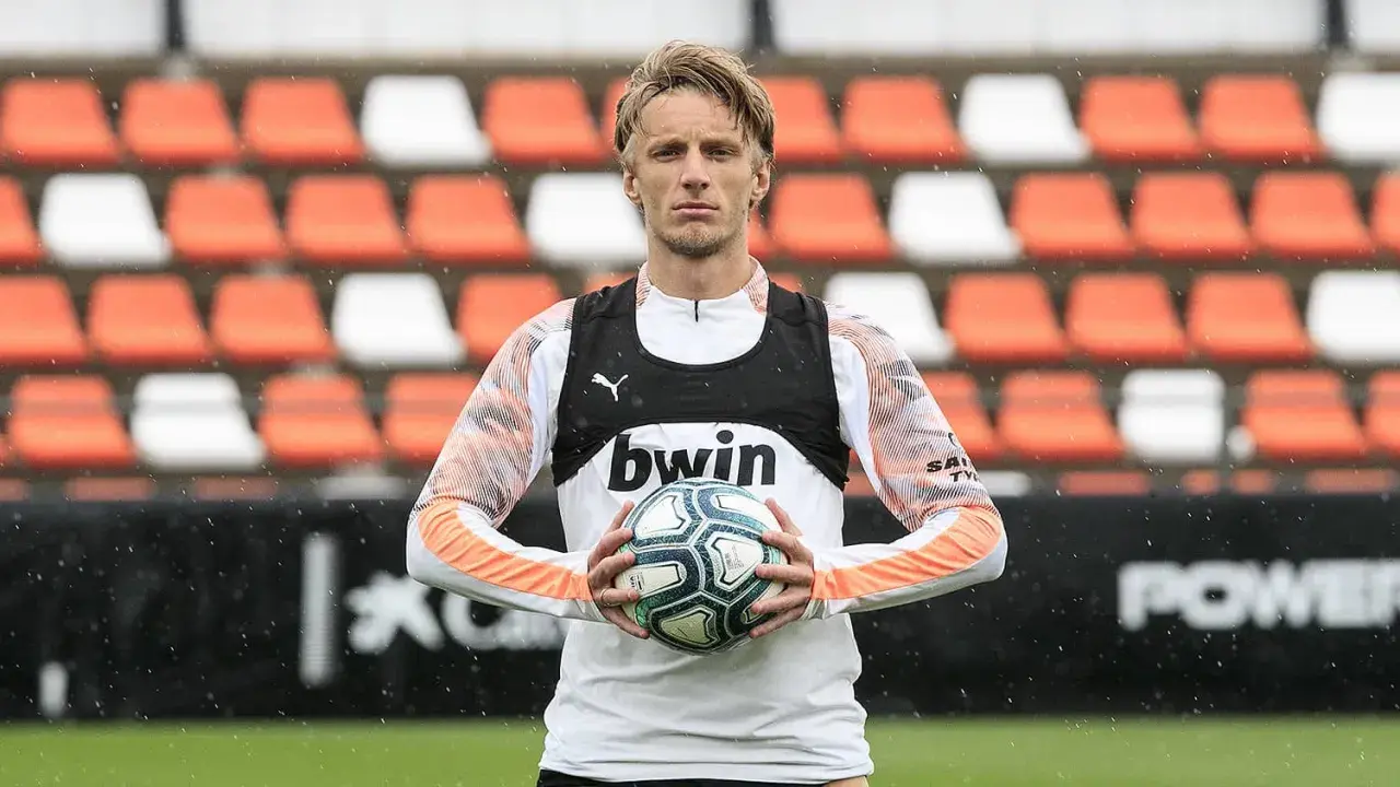 Piłkarz z blond włosami trzyma piłkę, ubrany w białą koszulkę z pomarańczowymi rękawami i czarną kamizelkę z logo bwin. W tle pomarańczowo-białe trybuny.