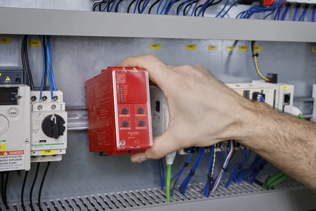 Ręka instalatora montuje czerwony sterownik bezpieczeństwa w szafie sterowniczej, wśród przewodów i innych modułów.