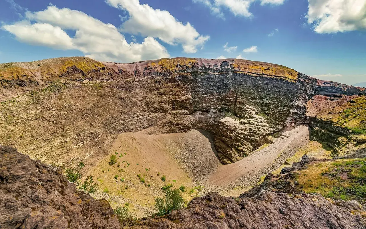 Il cratere del Vesuvio, con le sue pareti stratificate e il cono di detriti al centro, sotto un cielo azzurro con nuvole bianche.