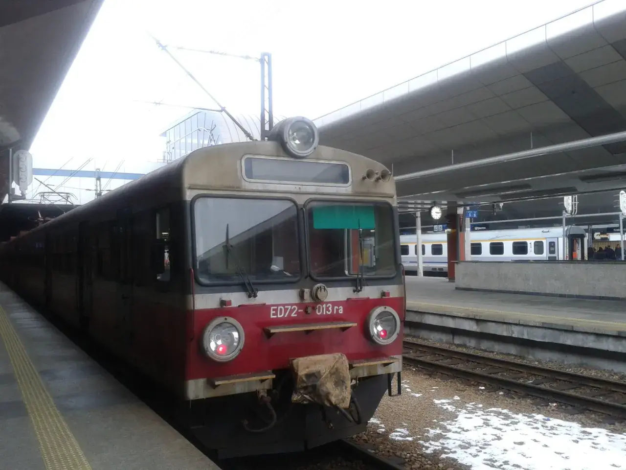 Pociąg ED72-013 ra stoi na peronie. W tle widać nowoczesny budynek dworca i inne pociągi.