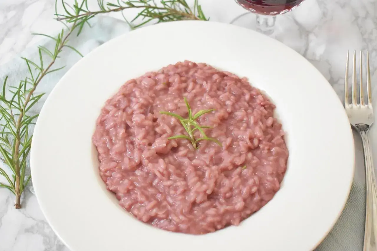 Aksamitne risotto z czerwonym winem, udekorowane gałązką rozmarynu, prezentuje się apetycznie na białym talerzu. Obok widoczny widelec i kieliszek wina.