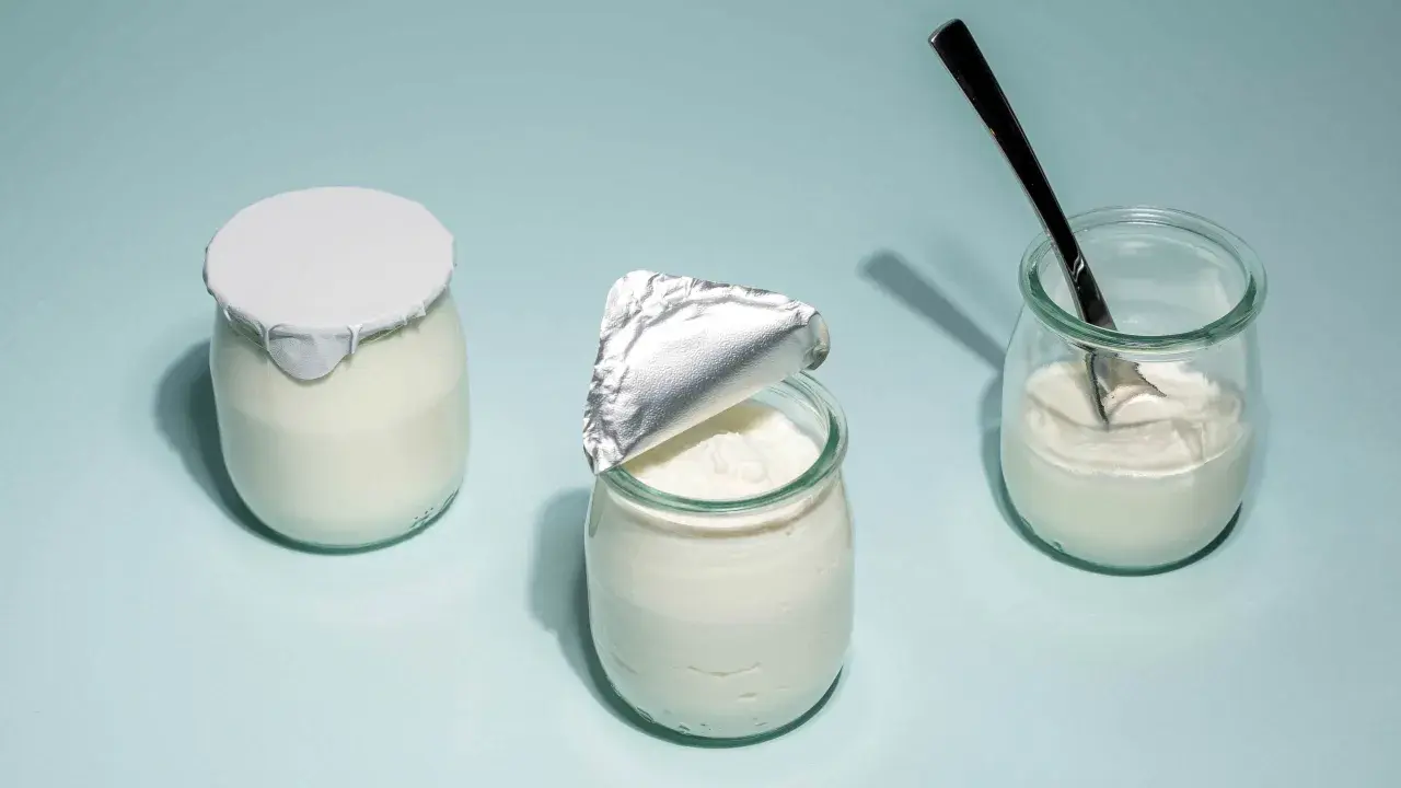 Tre burkar med hemmagjord yoghurt på ljusblå bakgrund. En är helt försluten, en annan är delvis öppnad och en tredje har en sked i sig.