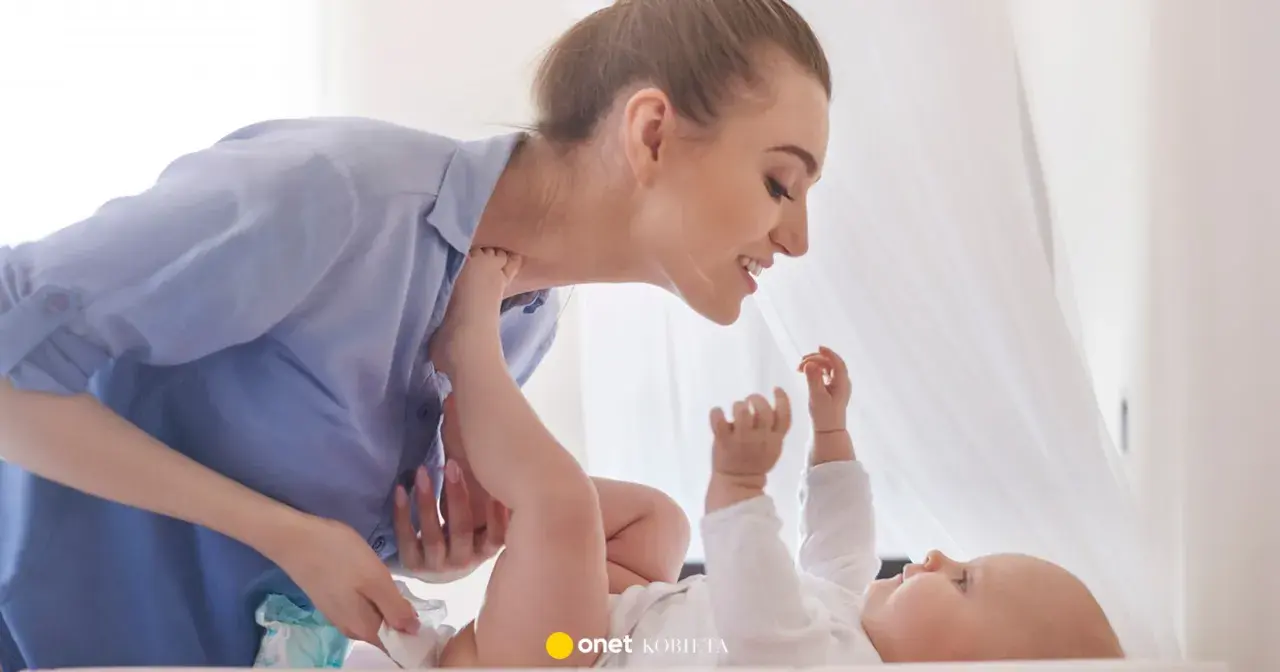 Uśmiechnięta mama pochyla się nad dzieckiem, które leży na plecach. Wykonuje szerokie pieluchowanie, delikatnie unosząc nóżki malucha.