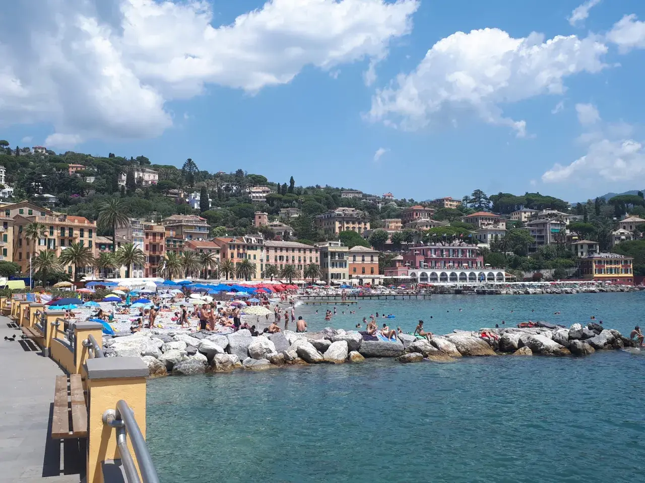 Plage animée à Santa Margherita Ligure, Italie, avec des parasols colorés, des baigneurs sur les rochers et des bâtiments pittoresques à flanc de colline.