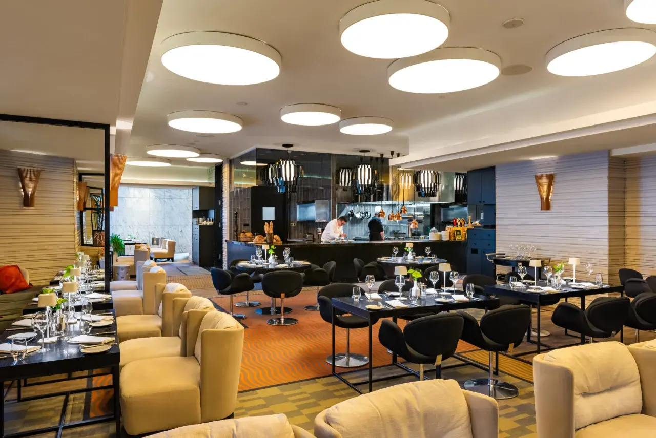 Elegancka brasserie z widokiem na kuchnię, gdzie szef kuchni przygotowuje dania. Stoliki nakryte do kolacji, wygodne fotele i nowoczesne oświetlenie tworzą przytulną atmosferę.