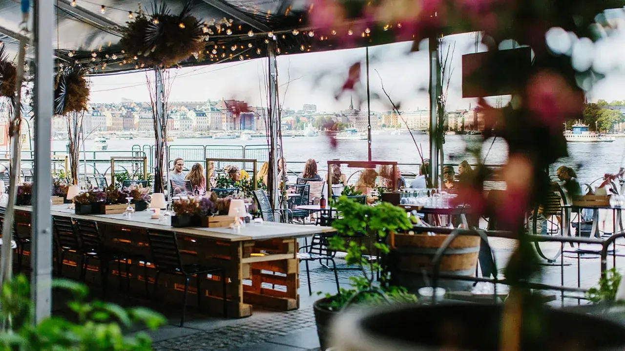 En mysig fotografisk veranda med utsikt över vattnet och stadens silhuett. Människor sitter vid borden och njuter av atmosfären.