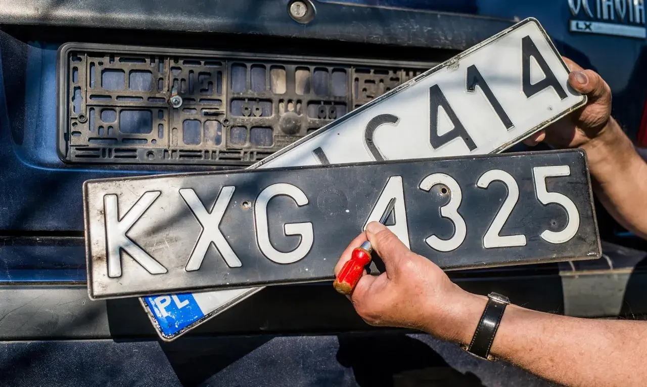 Wymiana tablic rejestracyjnych. Jedna tablica z numerem KXG 4325 jest przykręcana, druga z numerem C414 jest trzymana w ręku.