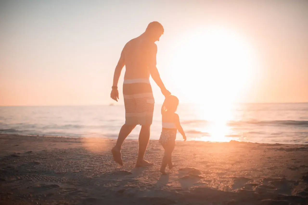 Ojciec prowadzi za rękę swoje dziecko 4 lata, spacerując po plaży o zachodzie słońca.