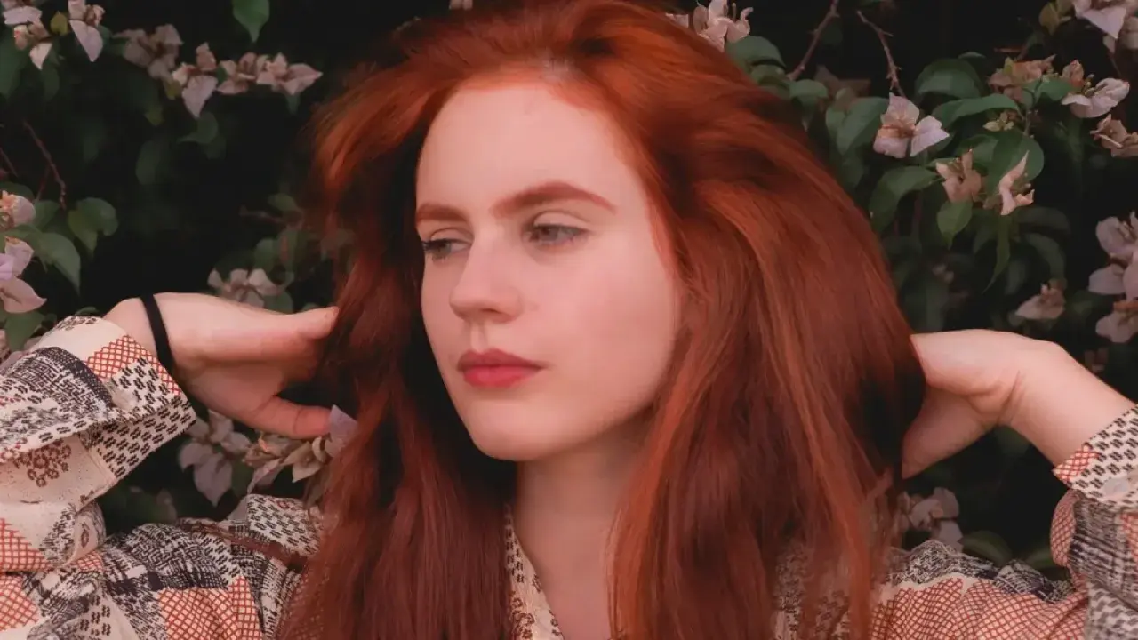 Chica con vibrante pelo pelirrojo, rodeada de flores y follaje, con las manos jugando con su cabello.